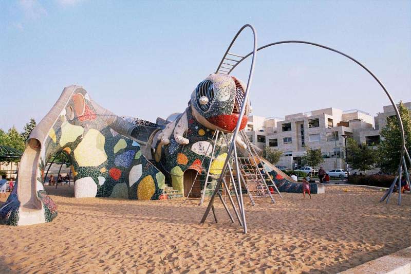 Красочное сооружение напоминающее кузнечика на детской площадке на фоне песка и зелени растений и неба 