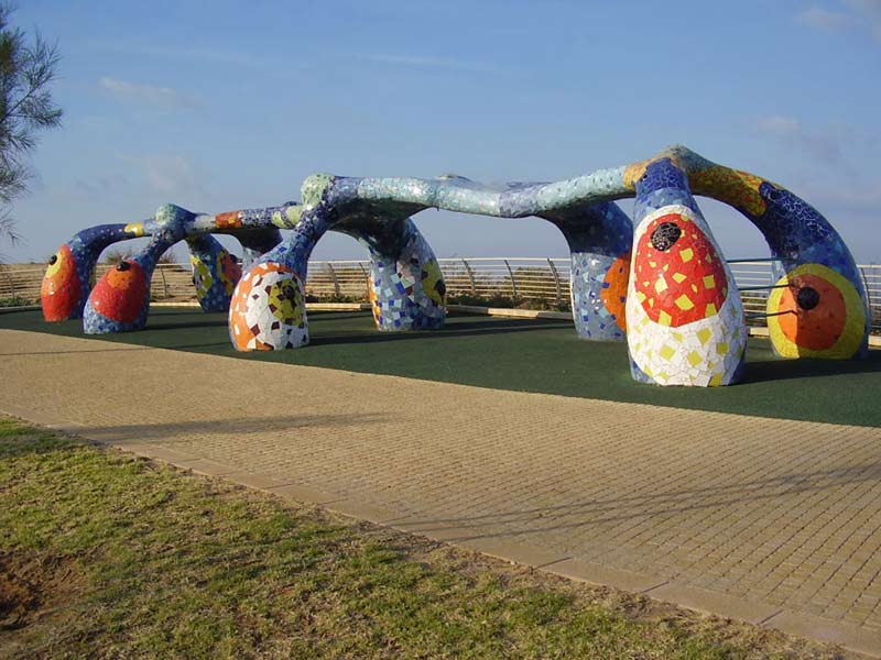 Пришелец с копытцами - скульптура Руслана Сергеева Длинный хребет с конечностями с утолщениями внизу основания из мозаики ярких радостных цветов на фоне зелёного покрытия и мощёной дорожки и зелени лужайки